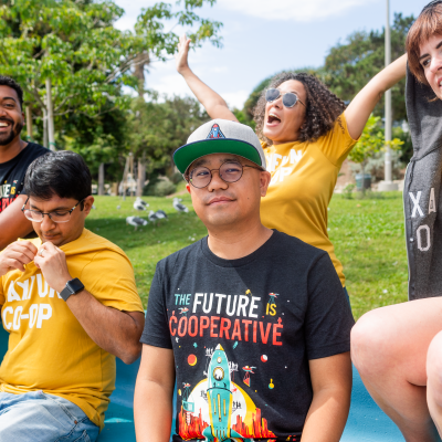 Five MaxFun worker-owners posed across a bench in a park wearing MaxFun co-op themed merch.